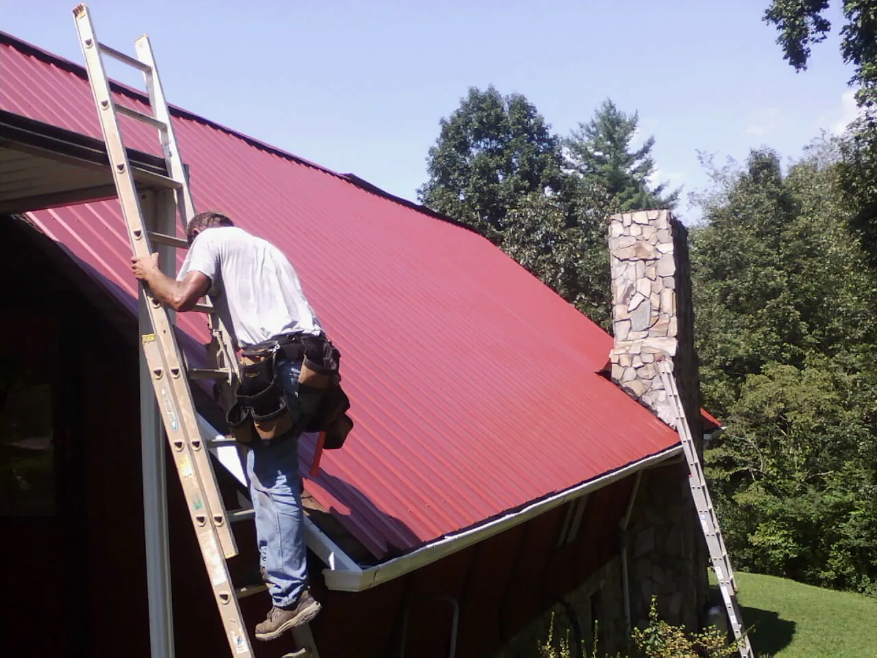 Roofer on a ladder beside a red metal roof and stone chimney — Soffit & Fascia Repair by Roofing Brothers in Sun River